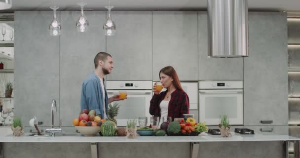 Frumos cuplu tânăr dimineața bea suc de portocale în bucătărie ei fericit zâmbind și au o masă plină de legume și fructe . — Videoclip de stoc