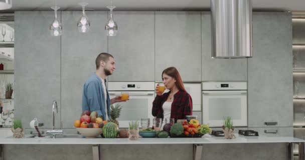 Belo jovem casal de manhã bebendo suco de laranja na cozinha eles feliz sorrindo e ter uma mesa cheia de legumes e frutas . — Vídeo de Stock