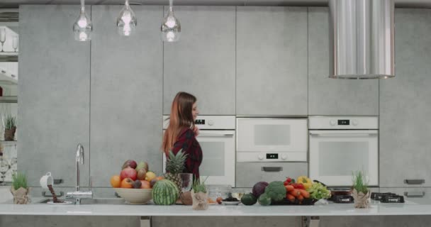 Mujer joven bailando en la cocina mientras hace el desayuno feliz moviéndose y haciendo cara divertida, sosteniendo una batidora de la mano, cocina gris moderna con una gran isla de cocina moderna. 4k — Vídeo de stock