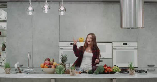 En una cocina moderna y hermosa con una gran isla de cocina, una hermosa joven bailando mientras hace comida y juega con un gran pimiento amarillo. — Vídeo de stock