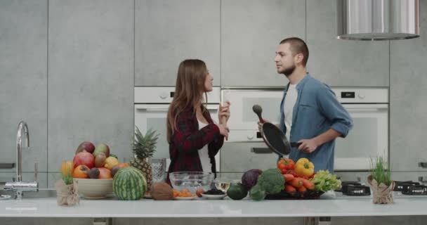 Dos carismáticos jovencita y chico dacing y cantando en la cocina mientras que la preparación de la comida, por la mañana, chico sosteniendo una sartén y la señora una mano mixer se mueven divertido, completa isla de la cocina son — Vídeos de Stock