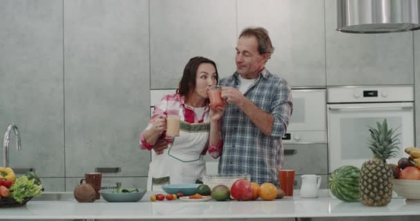 De manhã, um casal maduro saborear os smoothies de copos diferentes, eles têm um café da manhã saudável na parte da manhã em uma cozinha moderna . — Vídeo de Stock