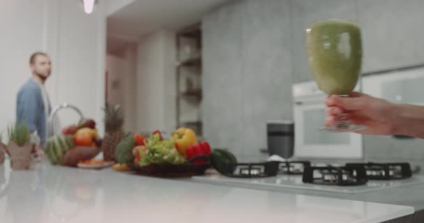 In de ochtend moderne keuken jonge gezin een gezonde groene smoothie, lady nemen het glas smoothie drinken en brengt aan haar partner. — Stockvideo