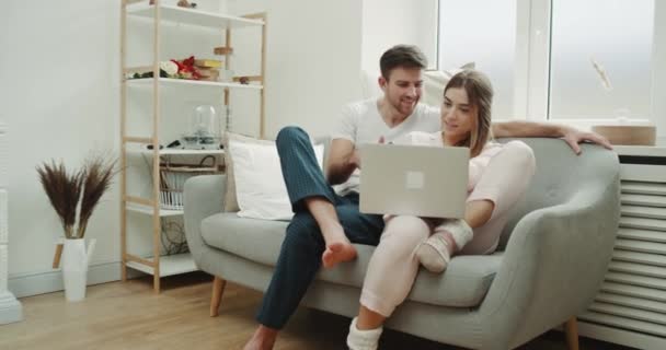 Утром пара в пижаме в стильной гостиной заказывает что-то из телефона, и, кстати, пользуется ноутбуком . — стоковое видео