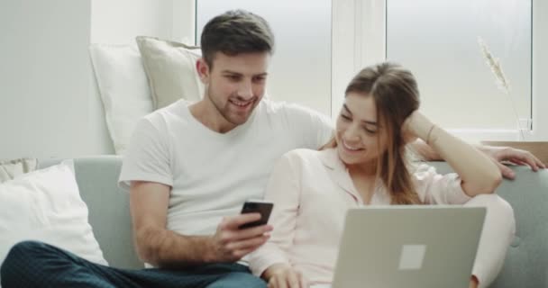 Casal de manhã fazendo uma ordem a partir do telefone e assistindo no caderno eles estão felizes sentados no sofá de pijama . — Vídeo de Stock