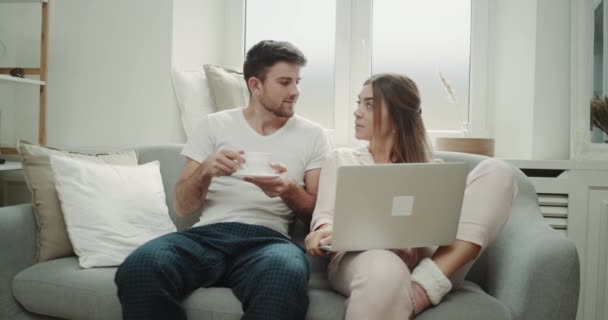 Mañana juntos para una pareja en pijama pasar tiempo juntos en el sofá hombre beber café dama viendo algo en el ordenador portátil, diseño moderno . — Vídeos de Stock