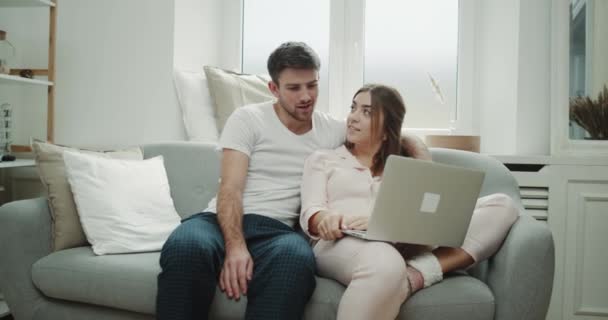 Pyjama's ochtend voor een paar in woonkamer zittend op de Bank, chatten en het gebruik van een laptop om iets. — Stockvideo