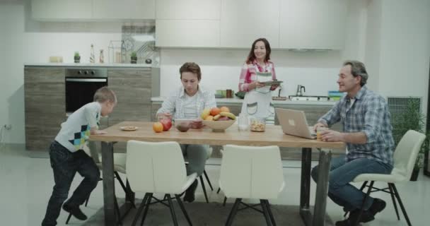 Attraktive Familienerwachsene morgens beim gemeinsamen gesunden Frühstück in der Küche, Papa hat seinen Laptop auf dem Küchentisch. Schuss auf rotes Epos — Stockvideo