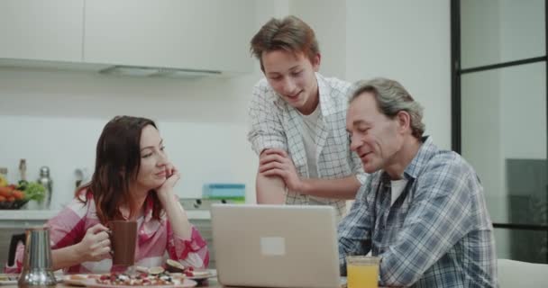 Son with his mature parents have a interesting conversation in the morning after the breakfast in the kitchen , while parents have a sit at dinner table , they watch something from the laptop. — Stock Video