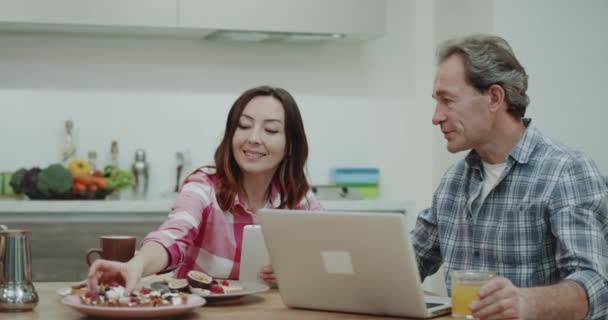 Frühstück zusammen in der Küche Reifes Paar isst ein gesundes Essen, Frau gibt ihrem Mann ein neues Rezept auszuprobieren, er arbeitet an seinem Laptop. — Stockvideo