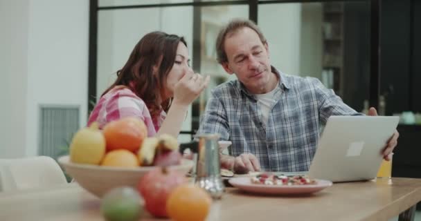 Charismatique couple d'âge mûr dans la cuisine, assis à table et regardant avec soin quelque chose sur l'ordinateur portable . — Video