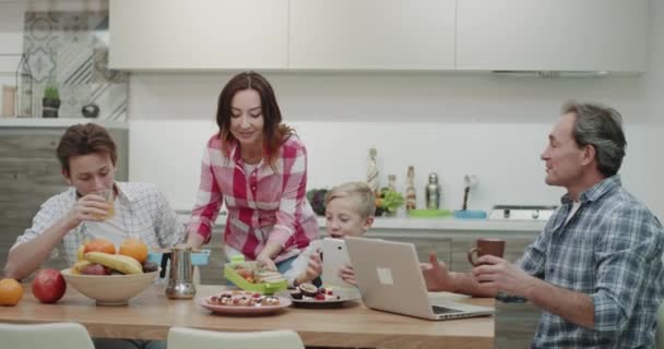 Morgens gibt die Mutter ihren beiden Söhnen die Brotdose und küsst sie glücklich, bevor sie zur Schule gehen Papa arbeitet an seinem Laptop. 4k — Stockvideo