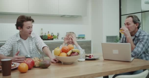 Привлекательная семья завтракает вместе со здоровой пищей, в удивительной кухне с бобовым дизайном, папа должен работать на своем ноутбуке, а сушить кофе мать поговорить со своими двумя мальчиками — стоковое видео