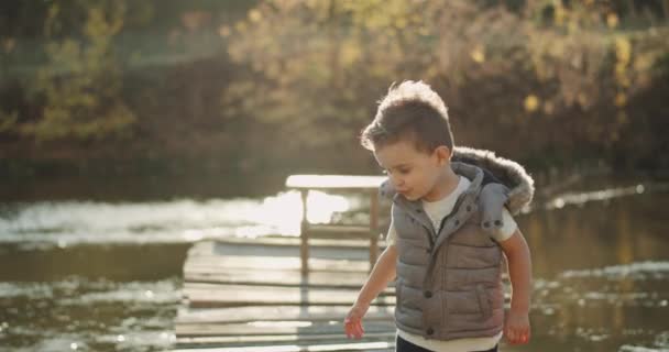 Retrato de un niño muy carismático de tres años de edad parado en el puente junto al lago y mirando directamente a la cámara mostrando un gran como — Vídeo de stock