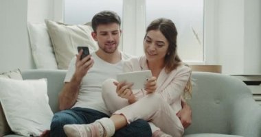 Bir tablet çift sıra--dan bir şey onlar kanepede oturan pijama ve mutlu bir ruh hali var telefon kullanarak.