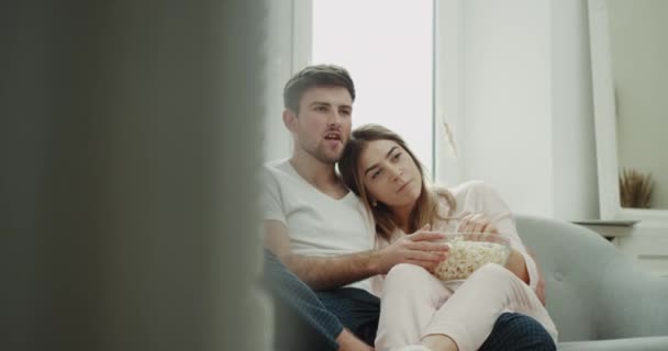 Romantisk tid Kramande par i pyjamas sitter i soffan och tittar på tv äta popcorn och glad utgifterna morgon tillsammans. — Stockvideo