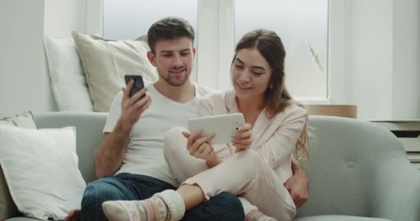 Використовуючи планшетну пару замовити щось з телефону вони знаходяться в піжамі, сидячи на дивані і мають щасливий настрій . — стокове відео