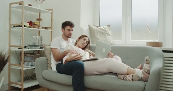 Lat dag för ett ungt par äta popcorn i soffan och titta igenom anteckningsboken klädd i pyjamas. — Stockvideo