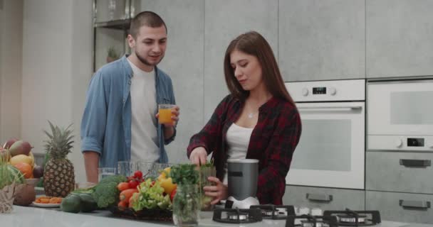 早上为一对年轻夫妇准备健康早餐, 男人喝橙汁, 女士在现代化的厨房里准备冰沙 — 图库视频影像