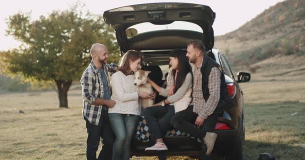Dos parejas tienen un viaje junto con un coche, tienen una parada sentado en el maletero del coche con un perro husky pequeño increíble y un niño pequeño en la parte posterior . — Vídeos de Stock