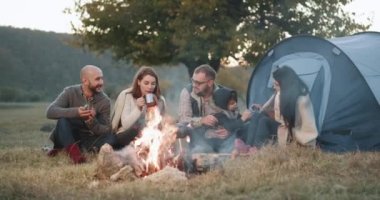 Bir grup arkadaşıyla birlikte, piknik çadır ile ve onların çocukla birlikte akşam oynayan genç bir aile kamp ateşi zamanı. 4k