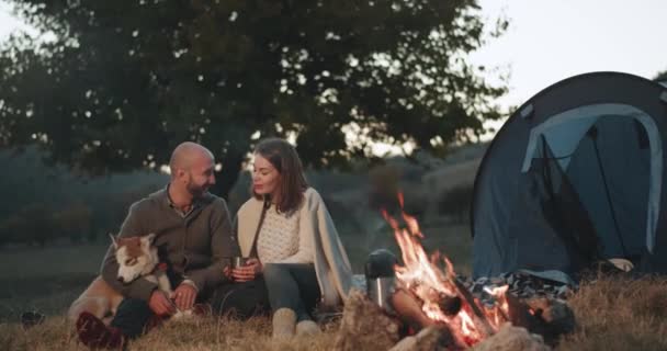 Hermoso vídeo de captura de dos jóvenes y el hombre tienen un momento romántico en el picnic en la noche al lado de la fogata que beben té y jugar con su perro husky. 4k — Vídeo de stock