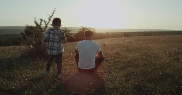 爸爸和儿子在大自然中度过了一段的时光 , 爸爸坐在草地上的小男孩从后面来到爸爸身边 , 亲吻着他那么可爱的风景. — 图库视频影像
