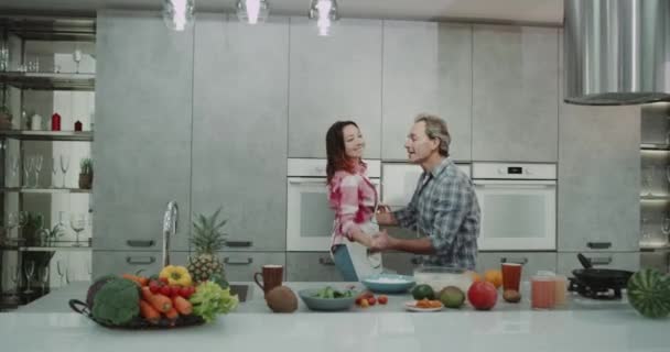 Schönes reifes Paar, das in der Küche tanzt, während es gesundes Abendessen kocht, modernes Design der Küche, große Kücheninsel. — Stockvideo