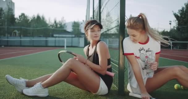 Dos chicas guapas después de un partido de tenis sentadas en el suelo y tomando una botella de agua . — Vídeos de Stock