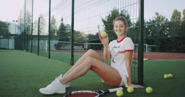 金髪キュートなレディ テニス コートで美しいテニス スーツを着て、床に座ってカメラの前でテニスのボールで遊んで. — ストック動画