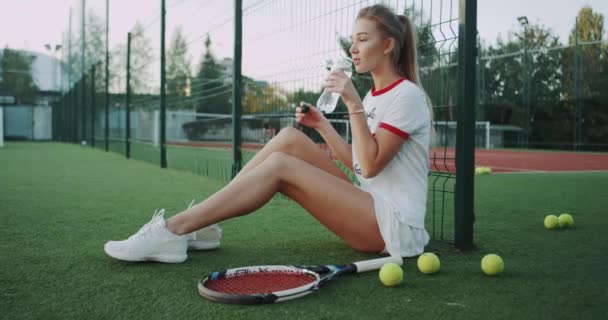 Beber botella de agua linda dama en la cancha de tenis al aire libre, sentado en el suelo, día soleado . — Vídeo de stock