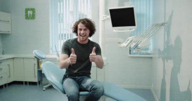 Diş hekimi sandalyede oturan ve onun sözlü onay için bekleyen bir dişçi Studio genç hasta adam modern bir diş kliniği kamera önünde büyük gülümseyen iyi bir ruh hali var. 4k