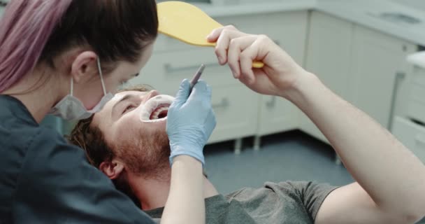 Primo piano giovane paziente carismatico e il suo dentista nella stanza dentale, dentista donna mostrando al suo paziente i risultati dopo una procedura di igiene orale, paziente con uno specchio per guardare ha un paradenti — Video Stock