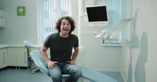 Zeer ontzagwekkende kerel met grote glimlach bezoek aan zijn tandarts in een medische kliniek zijn tanden selectievakje zittend op de stoel van de blauwe tandarts te wachten. 4k — Stockvideo