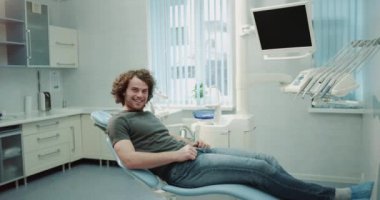 Büyük bir gülümseme ile kıvırcık saçlı Sevimli karizmatik adam onun dentis bir diş kliniği, o dentis sandalyede oturan ve onun doktor çok mutlu önünde gülümseyen için bekleyen bir randevu gerekecek