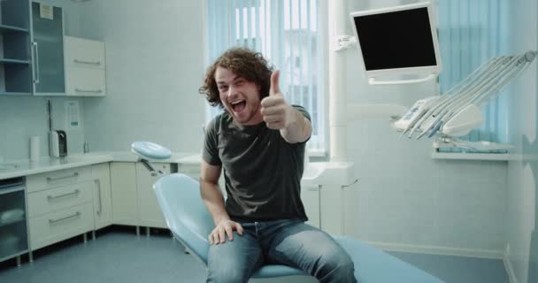 Hombre joven paciente muy carismático en una sala de la clínica dental sentado en la silla del dentis después de una comprobación de dientes muy feliz mirando delante de la cámara y mostrando un grande como. 4k — Vídeo de stock