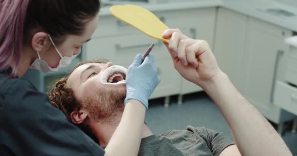 Moderna clinica odontoiatrica paziente con un paradenti dopo una procedura di igiene orale hanno uno sguardo utilizzando uno specchio per verificare i risultati che il dentista mostra al paziente il risultato. 4k — Video Stock