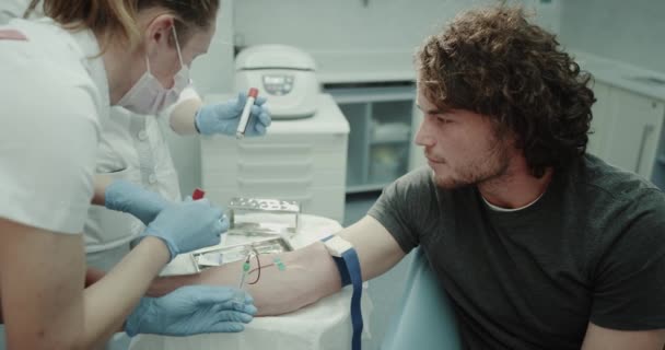 V moderní klinika asistent vzít krevní test z mladého muže, který sedí na židli, odběru krve z ruky. — Stock video