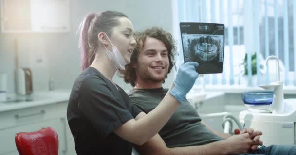 Dentist shows to his patient in a light dental clinic room teeth X-ray , and explain to him some details of his oral hygiene , patient are very happy. — Stock Video