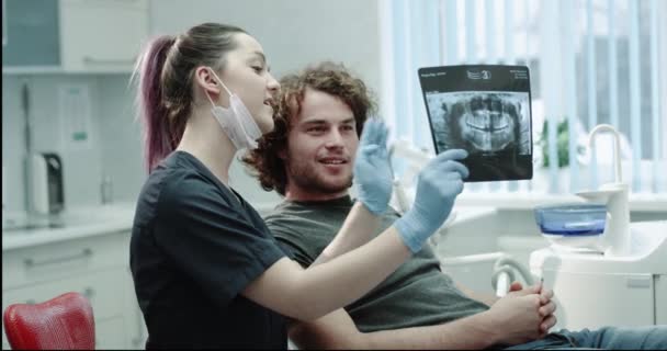 El paciente feliz en el dentista tiene una conversación amistosa con su médico, el dentista le muestra al paciente sus resultados de la radiografía de dientes, ambos sonríen en la habitación del dentista. tiro en épica roja — Vídeos de Stock