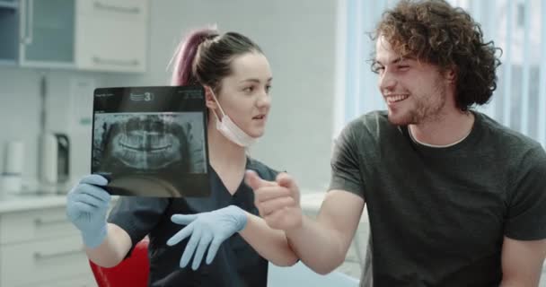 В стоматологической клинике молодые дантисты в синих перчатках объясняют результат рентгеновского снимка молодому человеку, у них отличная беседа и хорошее настроение, светлая стоматологическая комната . — стоковое видео
