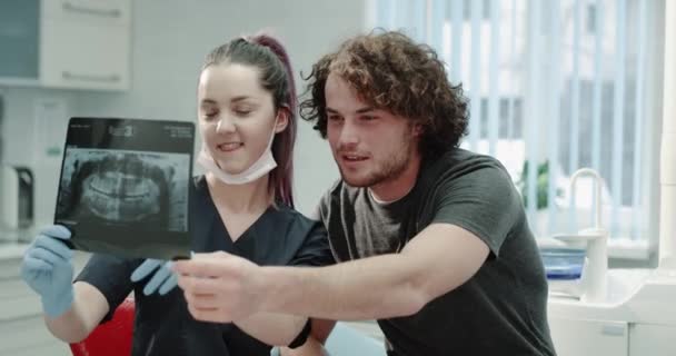 Ambiance très conviviale dans une salle de clinique dentaire dentiste expliquer soigneusement à son patient le résultat de ses dents rayons X, ils sourient, patient assis sur la chaise du dentiste . — Video