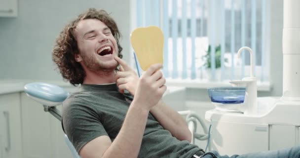 Ritratto di un giovane paziente dopo aver visitato il suo dentista, seduto sulla sedia blu del dentista e felice guardando attraverso lo specchio giallo ed esaminato i suoi risultati della procedura di igiene orale. 4k — Video Stock