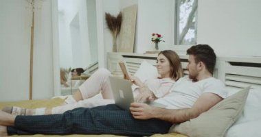 Pijama giyen ve bir kitap okuma ve laptop bir şey kontrol onların rutinleri yapmak yatakta genç bir çiftin güzel sabah.