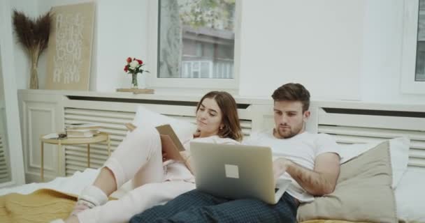 Rutina matutina para una pareja en la cama, señora leyendo un libro y hombre revisando algo en su cuaderno . — Vídeos de Stock