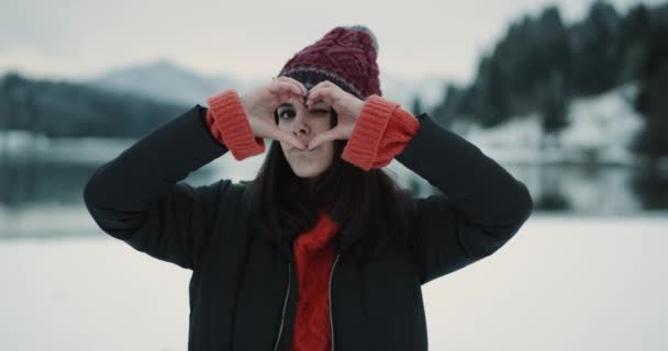 Portret van een mooie jonge dame toerist in het midden van een prachtig uitzicht op de achtergrond met een besneeuwde bos en een grote blauwe meer, met een groot hart gebaren met haar handen in de voorkant van de — Stockvideo