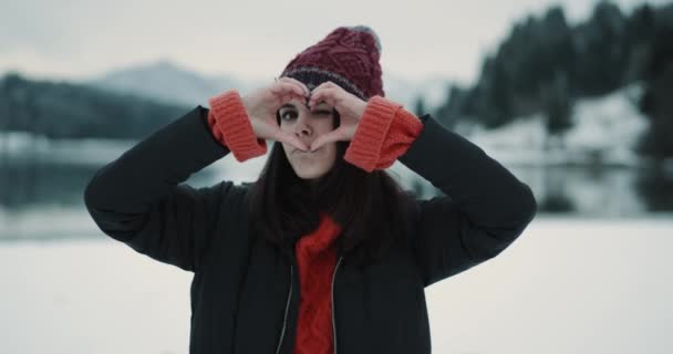 Retrato de una hermosa joven turista en medio de una vista increíble en el fondo con un bosque cubierto de nieve y un gran lago azul, mostrando un gran corazón gesticulando con sus manos frente a la — Vídeo de stock