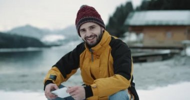Bir kış günü karizmatik genç turist turuncu ceket ve kırmızı şapka büyük kamerayı düz seyir ve bir büyük gösteren gülümseyen bir mavi closeup göl kıyısı yanında oturan mutlu