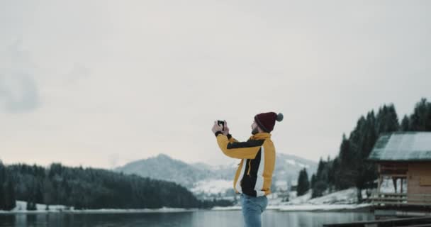 En medio de la naturaleza con un gran lago y la montaña nevada y el bosque joven con chaqueta naranja tomando fotos del paisaje de la tableta. 4k — Vídeo de stock