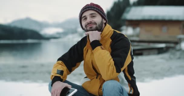 Portret van een jonge kerel zittend op een blauw boot in KITAmazing nature plaats met een besneeuwde meer en de bergen, op zoek direct naar de camera en glimlachend groot. — Stockvideo